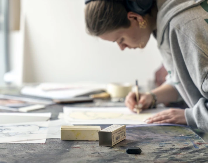 Person leaning over a table, drawing on paper in an art studio, with various sketches and art materials spread out Person leaning over a table, drawing on paper in an art studio, with various sketches and art materials spread out